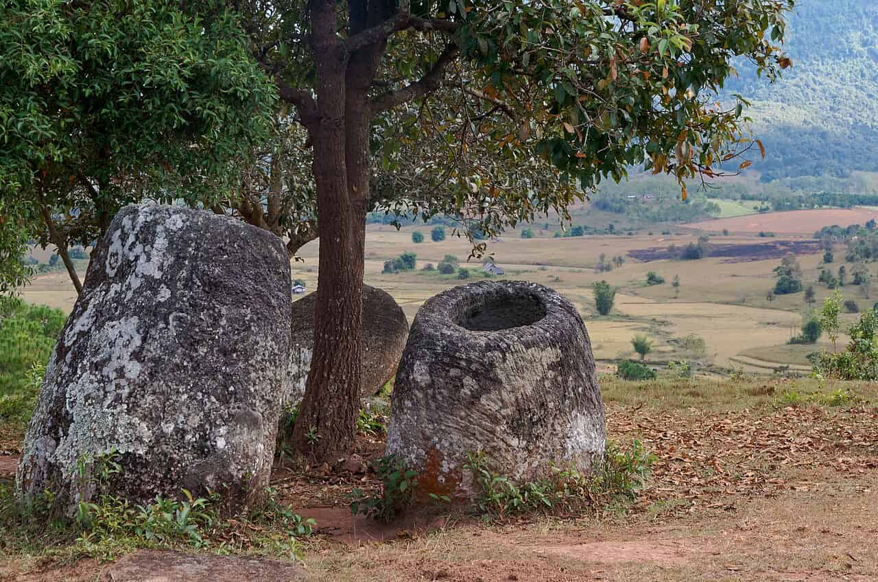 Two large ancient stone jars, roughly a metre tall, sitting on bare reddish-brown earth beneath a tree on a hillside. The jar on the right is upright with its hollow interior visible, while the one on the left is tilted. Both are weathered grey stone covered in patches of lichen. Behind them, a broad valley of farmland and scattered trees stretches into the distance, with green mountains on the horizon. Fallen leaves litter the ground around the jars.