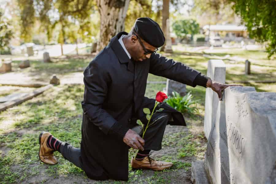 A gentleman holding a rose kneels before a gravestone.