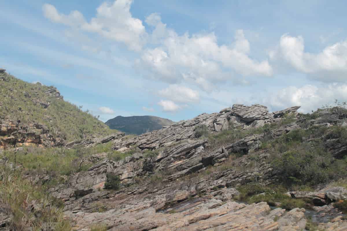 A rocky landscape, strewn with fractures and scrubby grasses. The land looks dry beneath the light blue cloud-speckled sky.