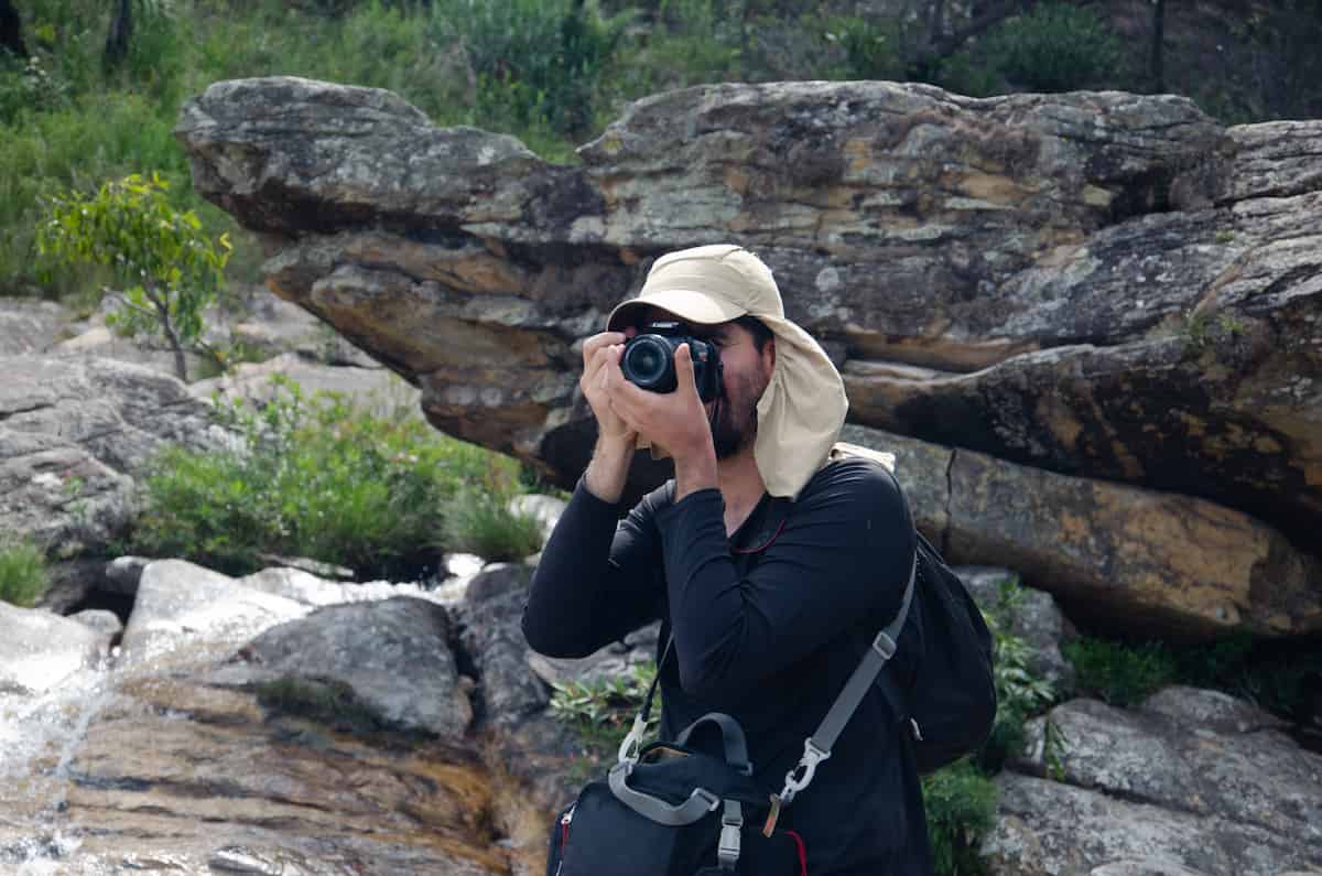 A handsome, bearded young man holds a camera to his eye to photograph some Science. Behind him are broken rock formations and tenacious small plants growing in crevices therein.