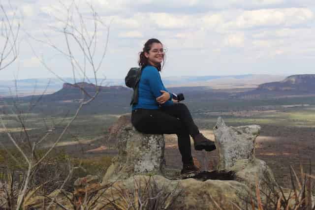 A hiker sits on a rocky outcrop overlooking an expansive valley landscape. The individual, wearing a blue top and carrying a backpack, is photographed in profile while seated comfortably on the natural rock formation. The background reveals a sweeping vista of rolling terrain and distant plateaus under a cloudy sky.
