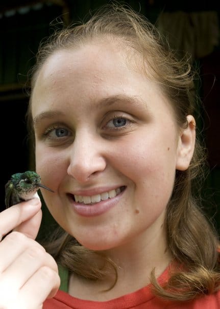 Dr. Laura Lagomarsino smiling at the camera while gently holding a small hummingbird in her hand during fieldwork in Costa Rica in 2008. The tiny bird has iridescent green plumage.