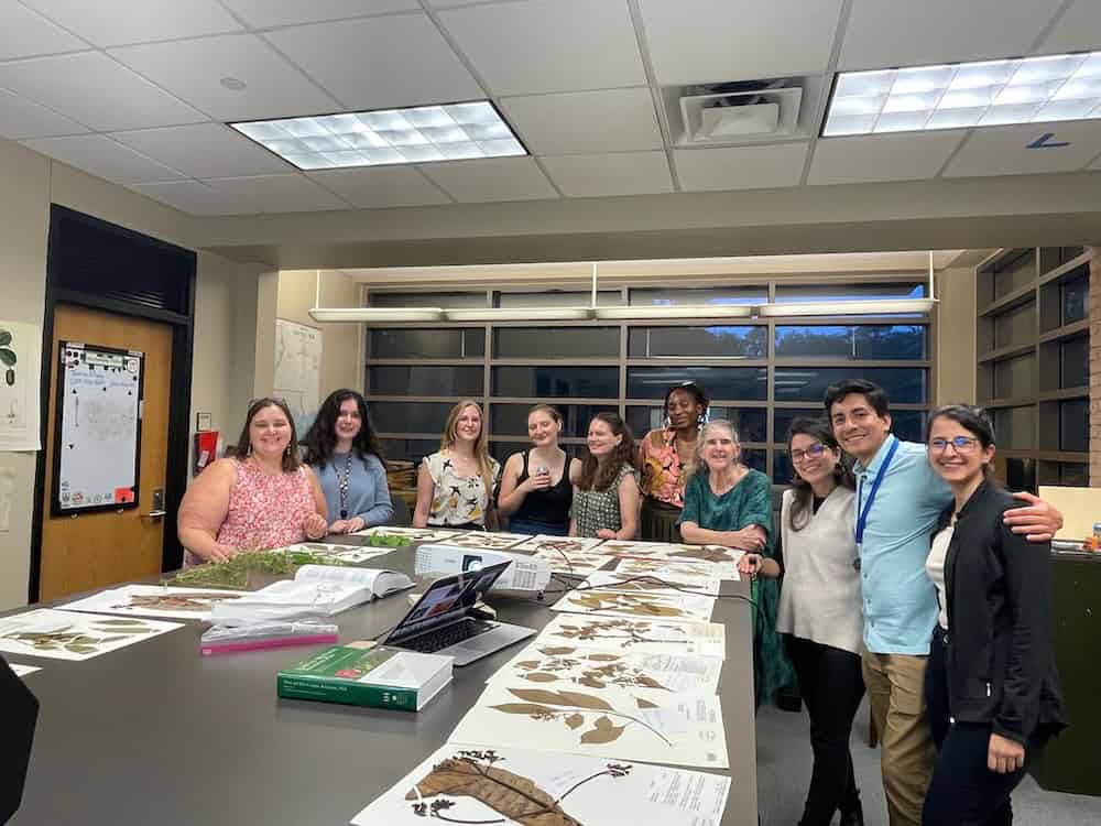 Dr. Laura Lagomarsino's research lab group in the Shirley C. Tucker Herbarium at Louisiana State University in April 2024. The group of approximately ten researchers stands around a large table covered with pressed plant specimens, herbarium sheets, and research materials. The herbarium setting features tall specimen storage cabinets in the background and fluorescent lighting typical of research facilities. 