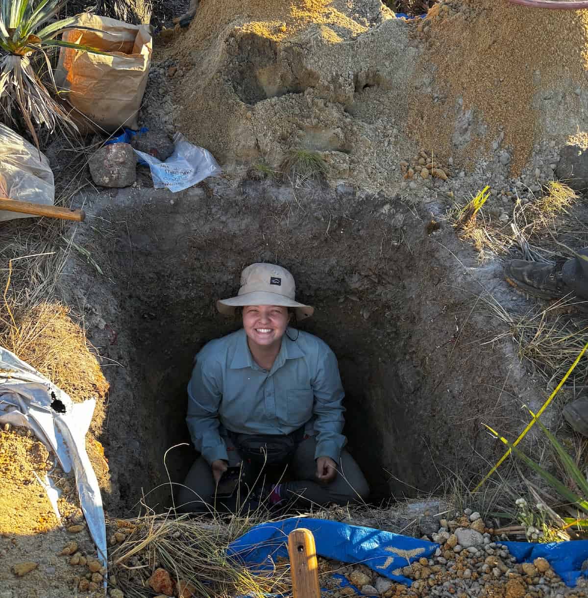 A woman in a soil pit looking up at a camera smiling.