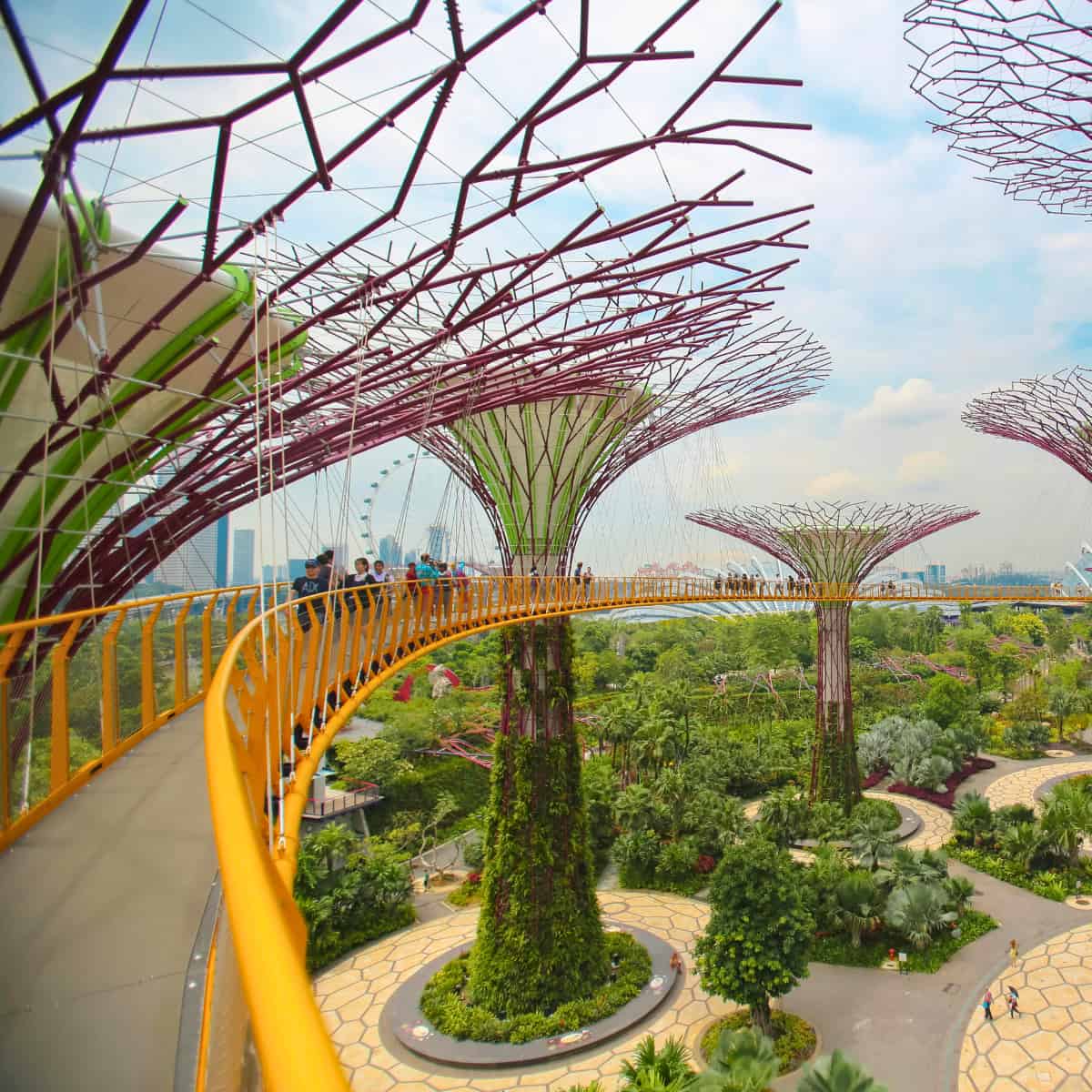An orange walkway passes high in the air through steel trees that, in their lower trunks are, covered by lush vegetation.