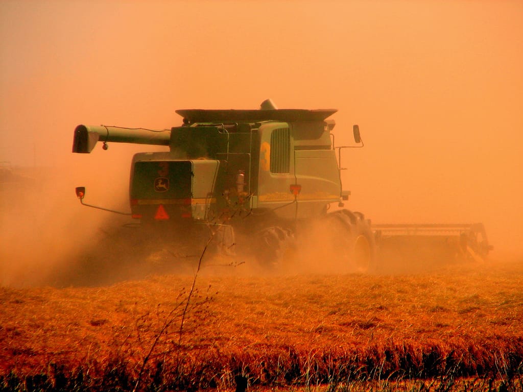 A rust red combine harvester running over rust coloured wheat against an orange sky.