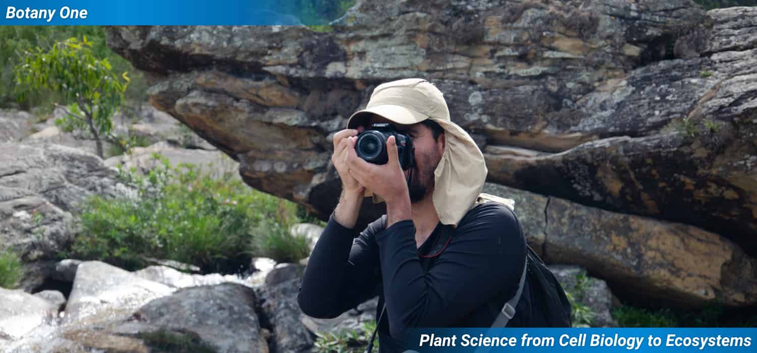 A young man points his camera to the horizon against a background of broken rocks and small thirsty plants.