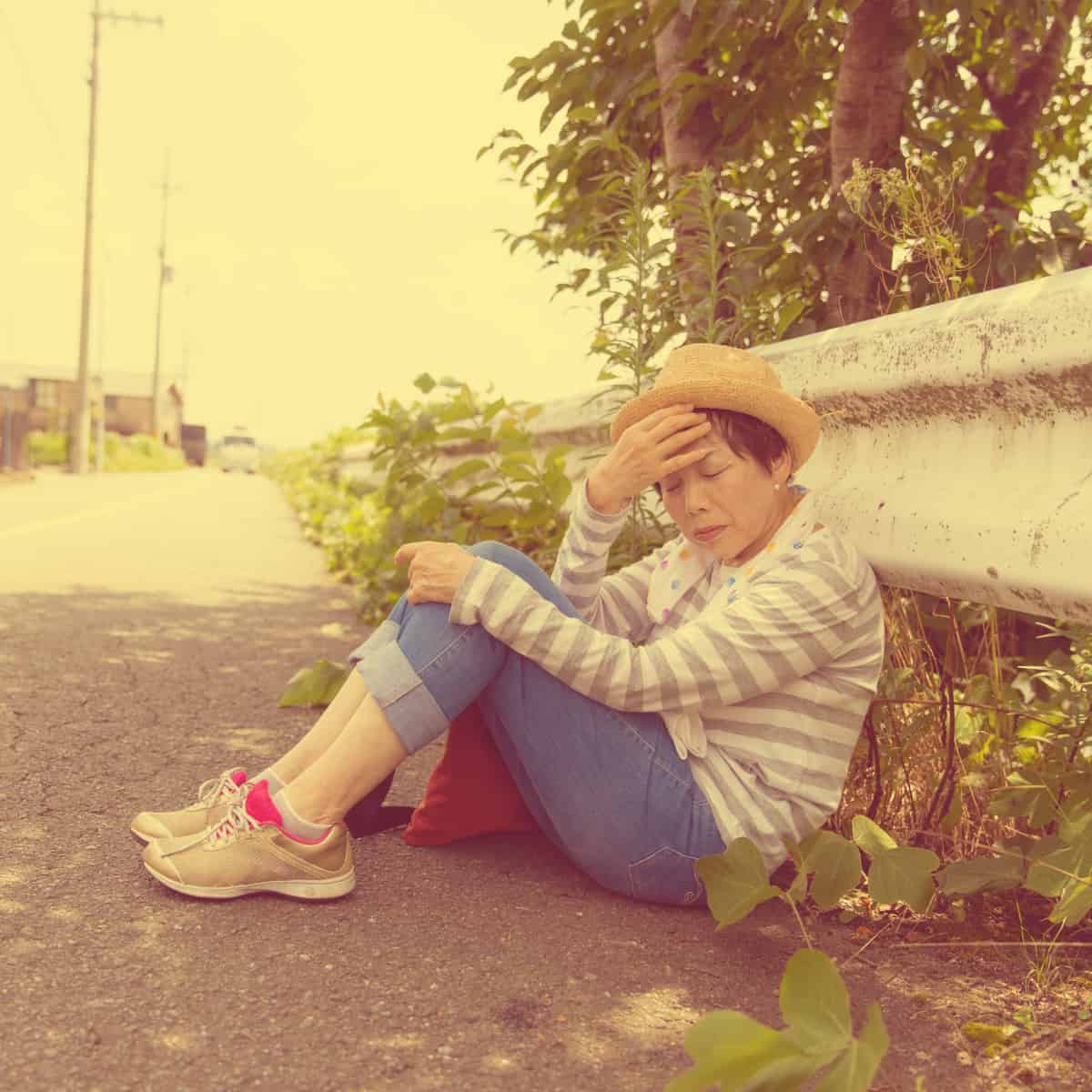A person in a striped shirt and hat sits on the ground leaning against a concrete wall, with one hand covering their face in