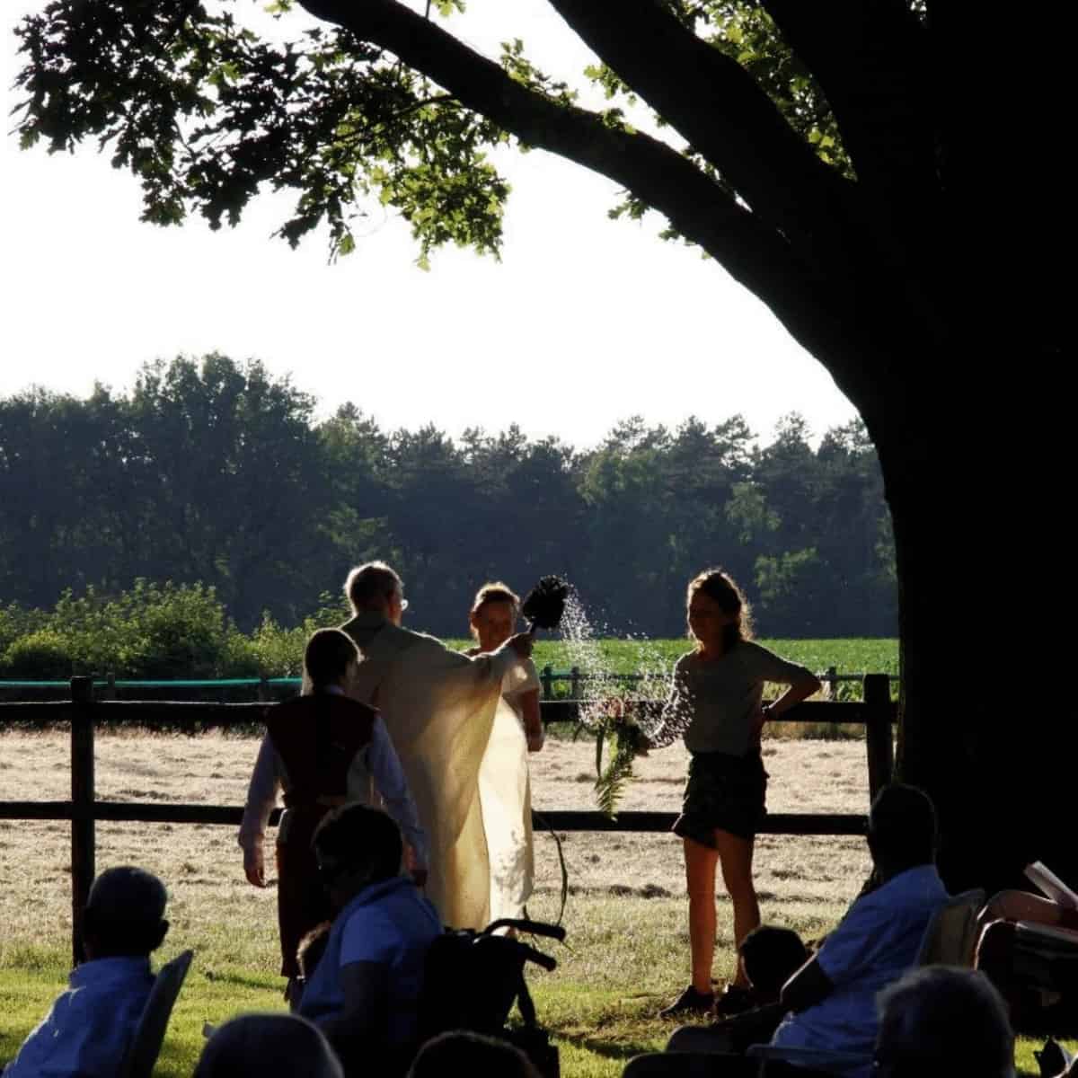 A young lady holds a wreath, while and elderly priest sprinkles water over it in the summer sunset. Other villagers can be se